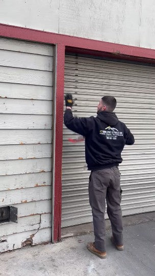 Person in a black jacket with a logo on the back, using a tool against a metal shutter.