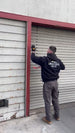 Person in a black jacket with a logo on the back, using a tool against a metal shutter.