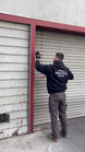 Person in a black jacket with a logo on the back, using a tool against a metal shutter.